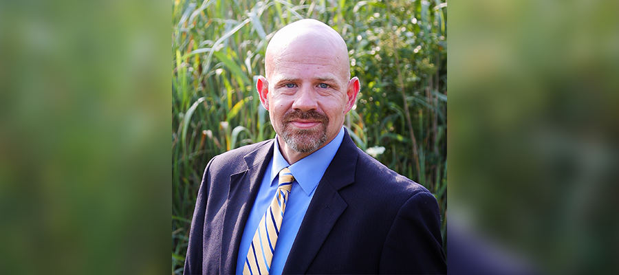 Rian Owens, Operations Manager at ACRT Pacific, wearing a suit and tie, standing outdoors with tall greenery in the background.